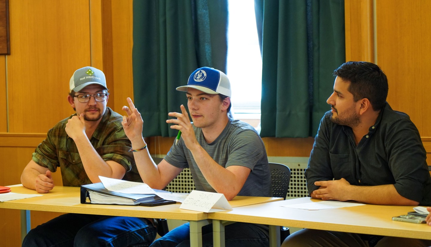 three men sit behind a table and one gestures with his hands as he talks