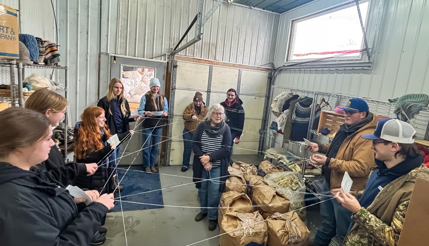 youth stand in a circle holding pieces of criss-crossed yarn while a woman stands in the midst of the yarn, talking