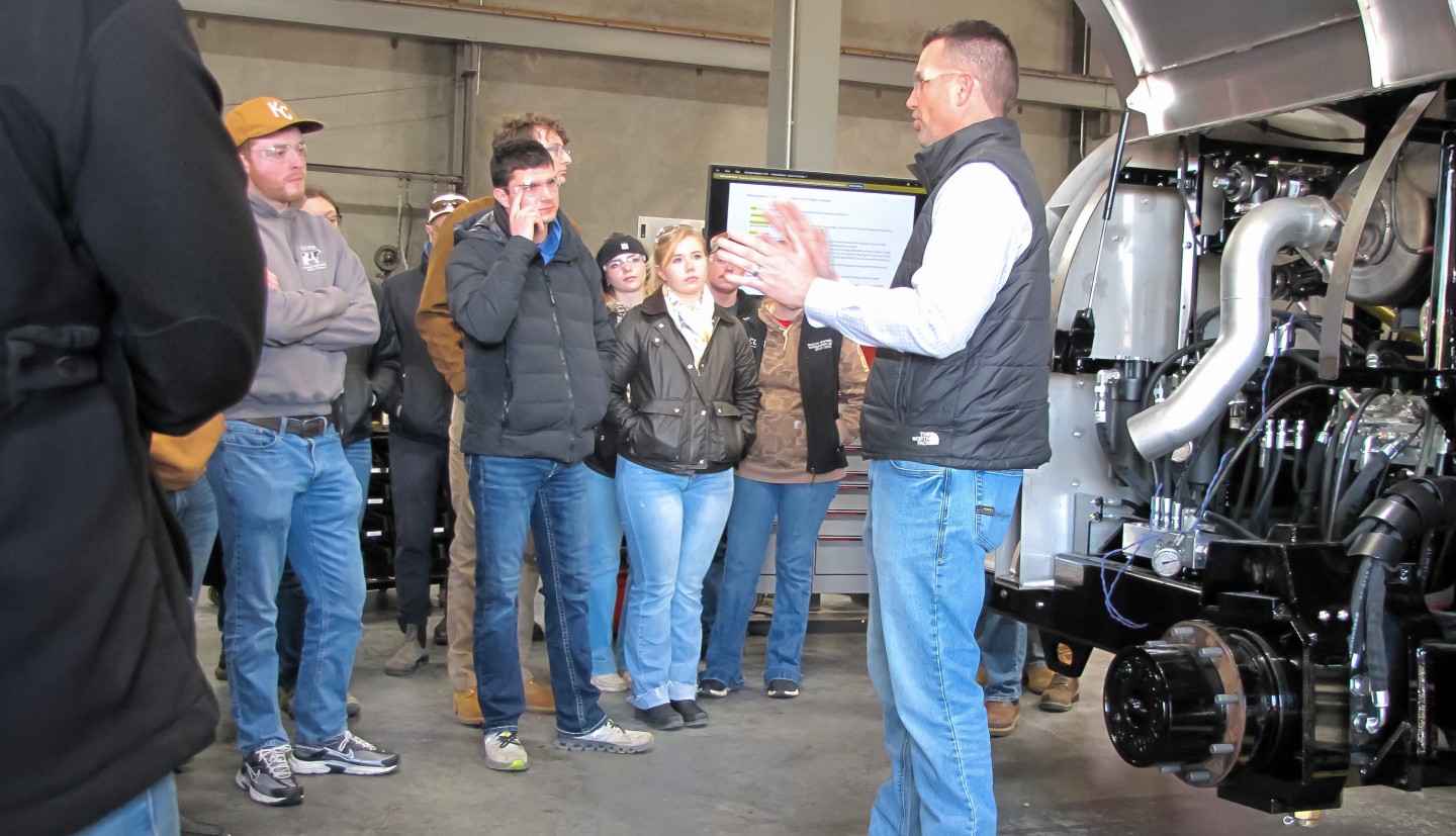 a man stands by some agricultural equipment and talks to a group of people