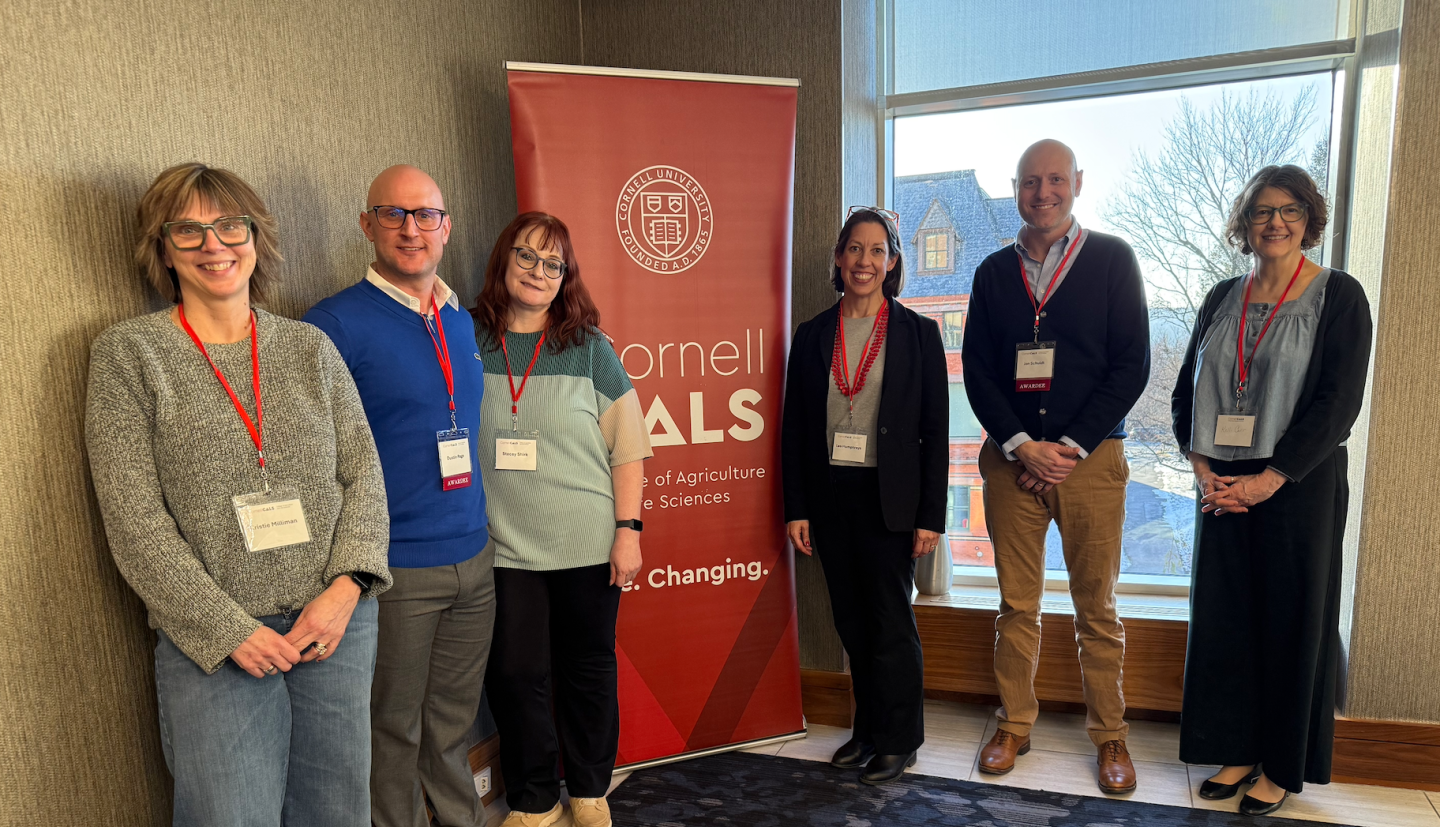 6 individuals stand and smile in front of Cornell CALS poster