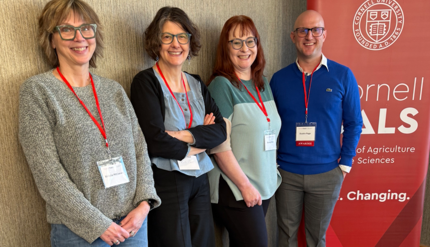 4 Cornell Comm staff members pose together in front of Cornell CALS poster. 