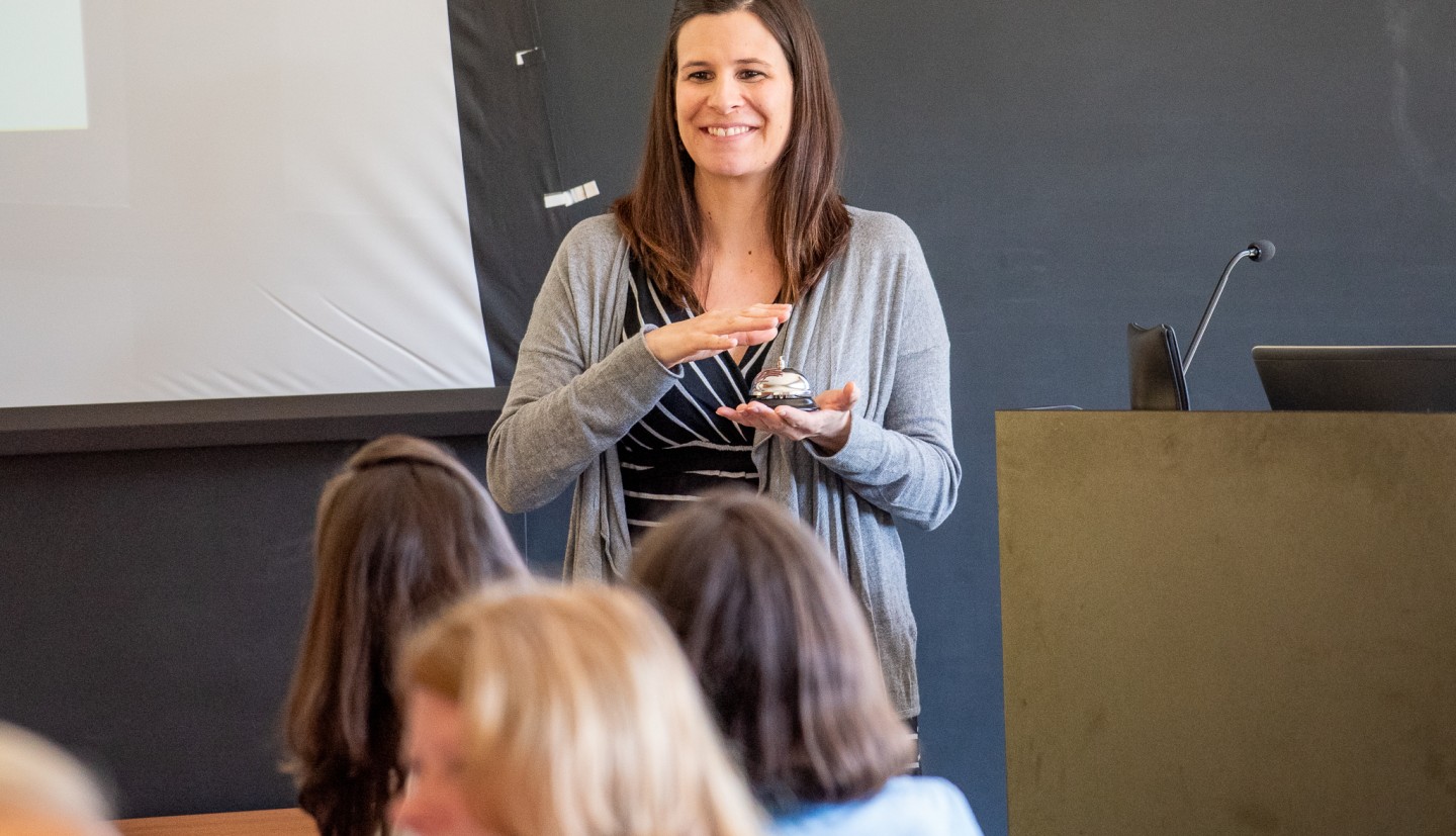 Cheryl in a classroom. He hand is going to ring a bell, for an activity connected to a nutrition education activity for kids.