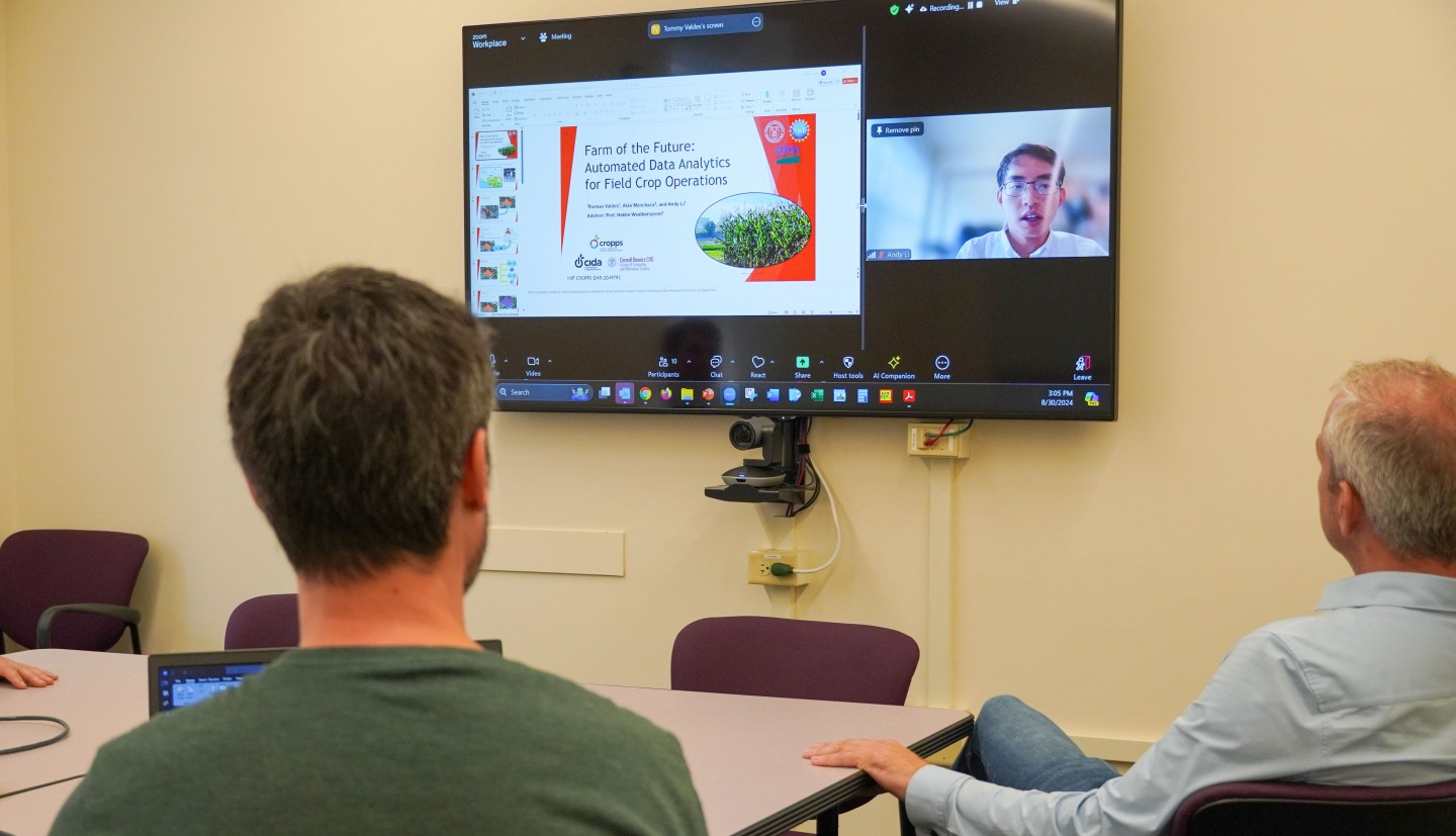 a screen on a wall shows data and the face of a man who is presenting by zoom while a couple of men sit facing the screen