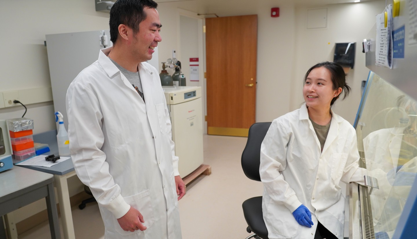 a standing man and a sitting woman in lab coats talk together in a lab