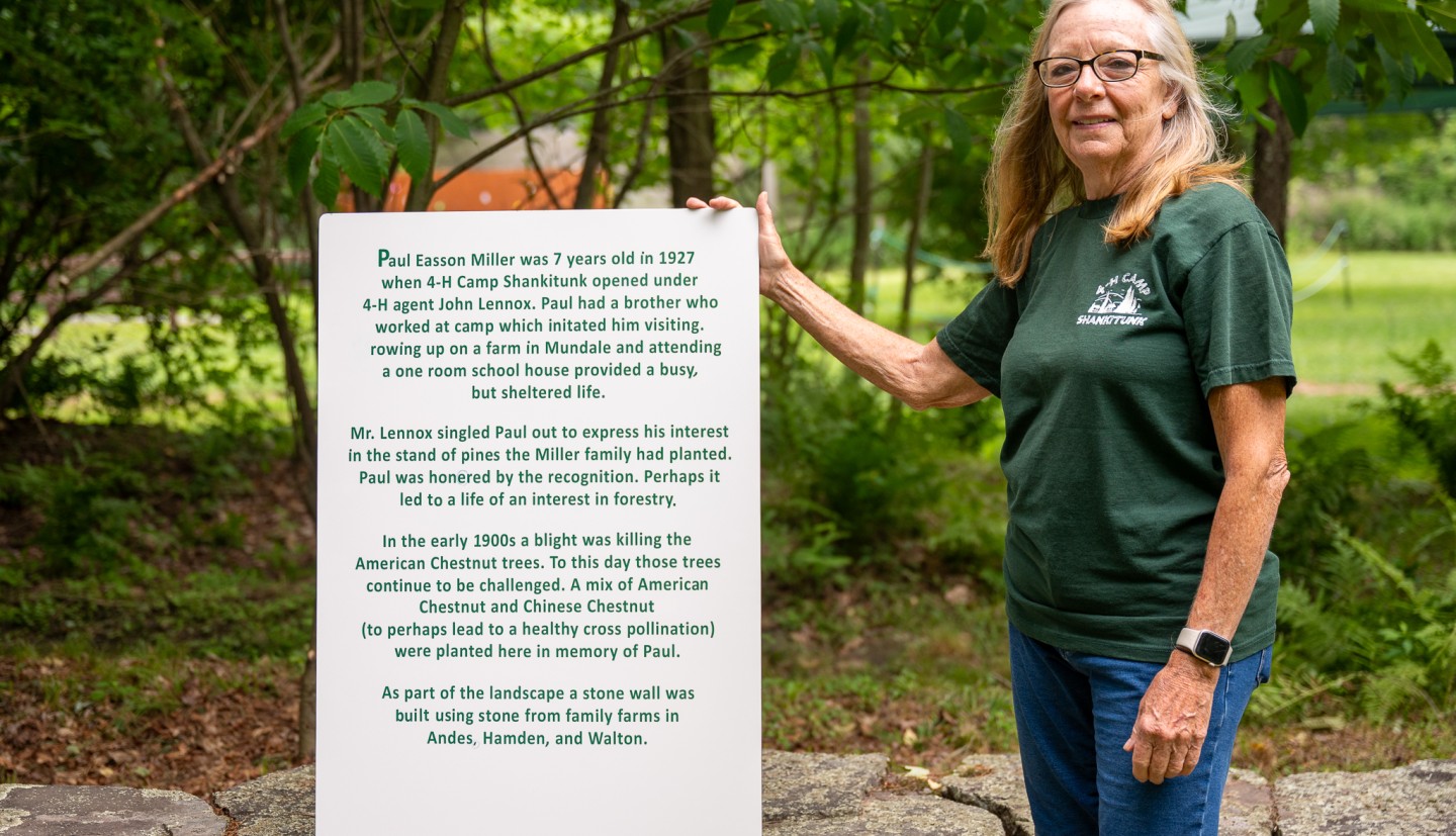 Lindy Tggart, 4-H volunteer, at Camp Shankitunk with a sign commerorating her fathers tree planting efforts