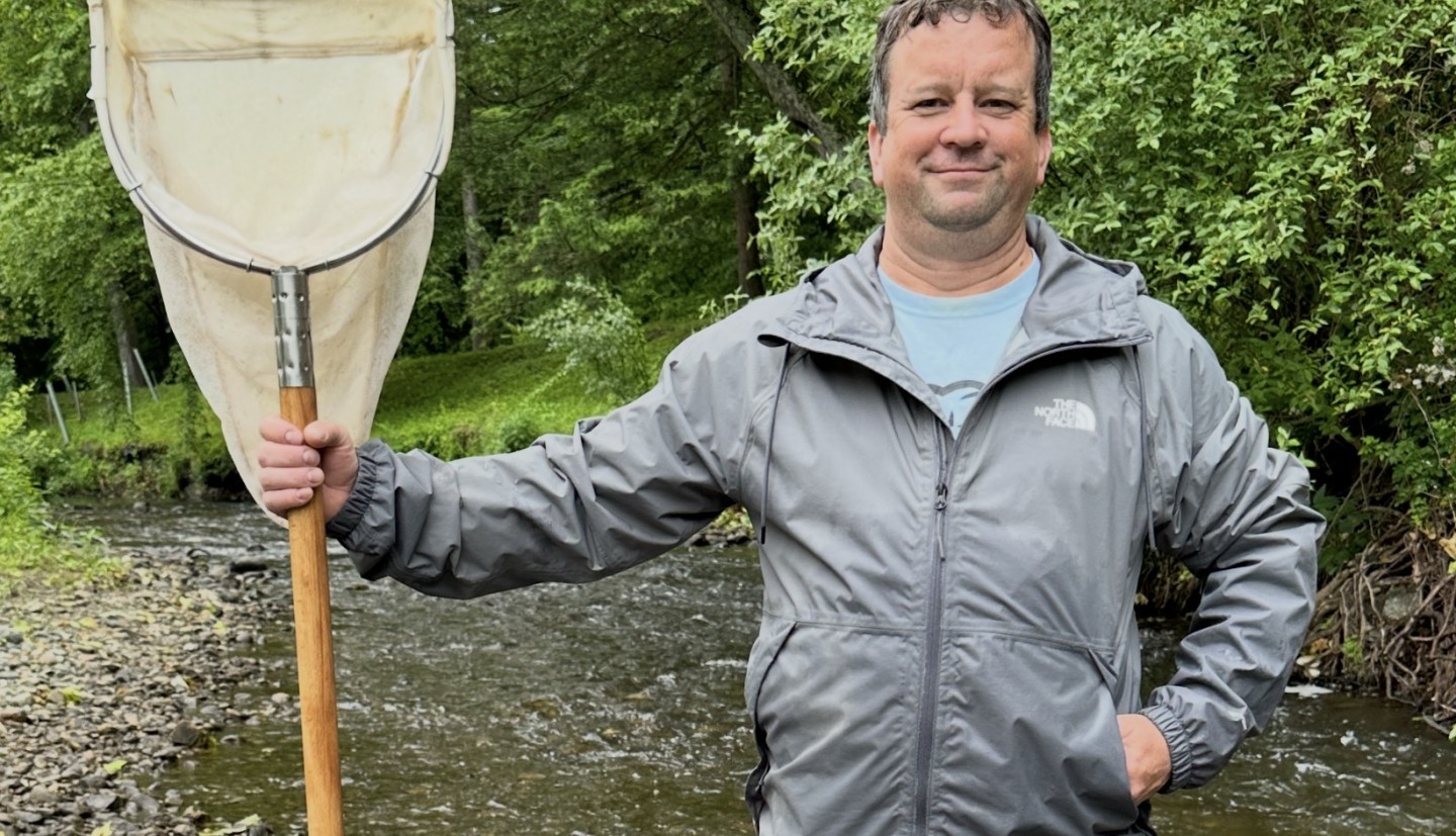 Photograph of a man in a rain jacket holding a large net standing in a stream.