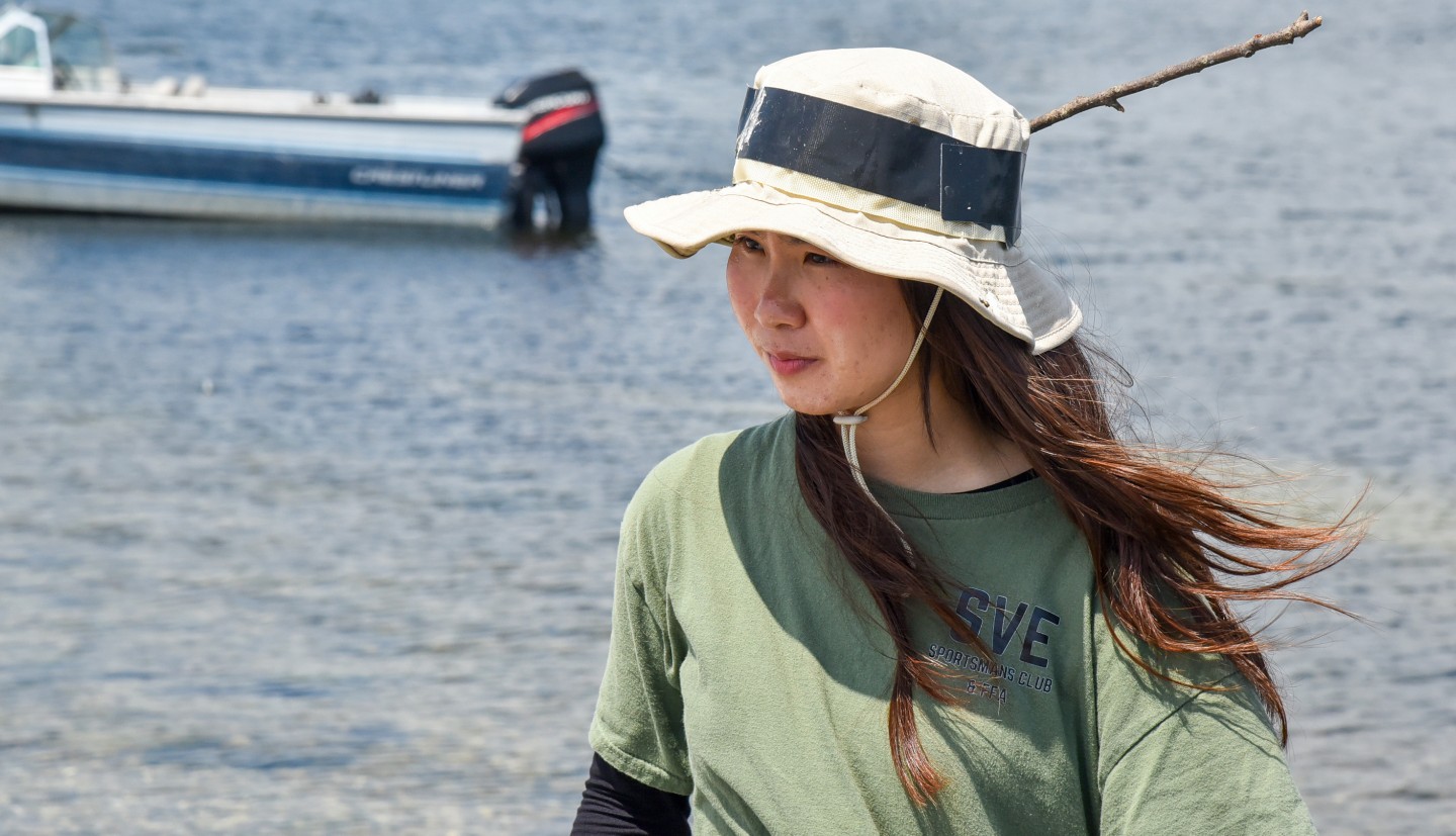 a woman wearing a sun hat walks with the wind blowing back her long hair and water and a boat in the distance behind her