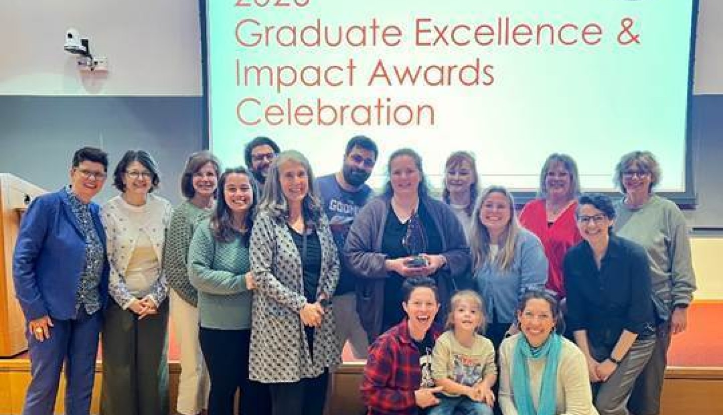 Joanna Alario holding an award at the Graduate Excellence and Impact Awards Celebration with a group of other people around her