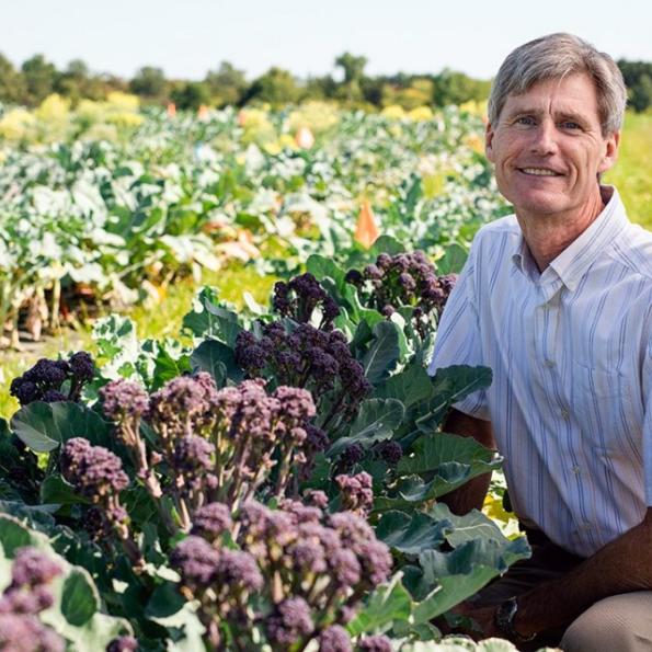 A white male crouching next to a broccoli plant and smiling in the middle of a large crop field of broccoli