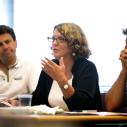 An older woman with grey hair sitting at a table with other adults, speaking with them and gesturing.