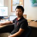 An asian male sitting at a desk in his office with a computer behind him