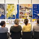 Several men and women sitting with their back to the camera looking at colorful posters of landscape architecture with a young woman in the front of the room presenting the posters