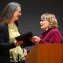 Two women facing each other standing on a stage in front of a podium with an award being passed off between them.  