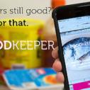 A hand holds an iPhone with the Foodkeeper application open on it in front of an ope fridge with containers of food in it