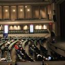 A female faculty member standing on a stage in a large performance hall to students scattered in the chairs below