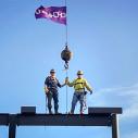 Two men stand on a construction scaffold flying a purple flag