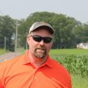 Photo of Andy Miller in an orange shirt and sunglasses, smiling with the farm in the background.