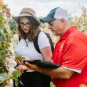 a woman in a hat and a man in a red shirt stand in a vineyard