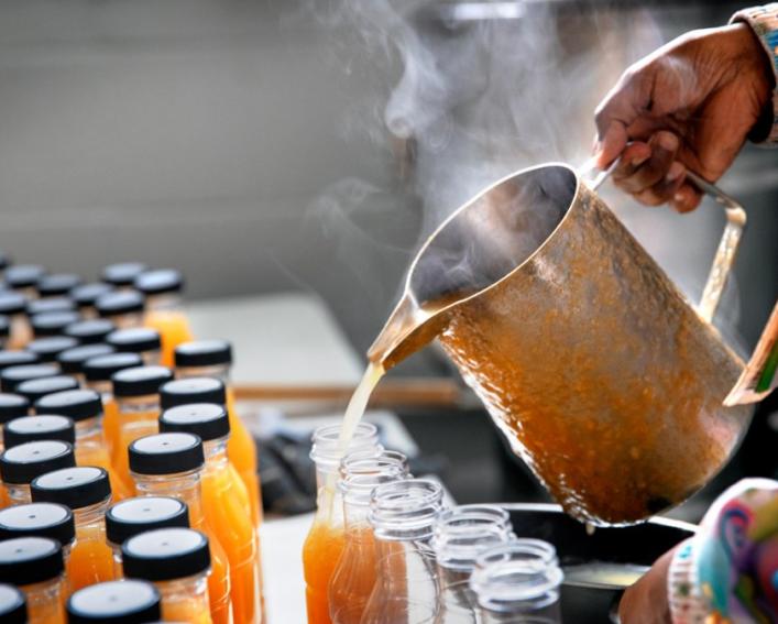 A hand holding a steaming, copper decanter that is pouring orange colored juice into bottles 