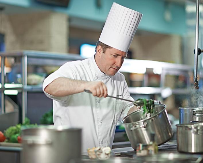 A male chef wearing a tall, white chef hat and white chef jacket spooning green beans out of a pot on a stove in a large kitchen 