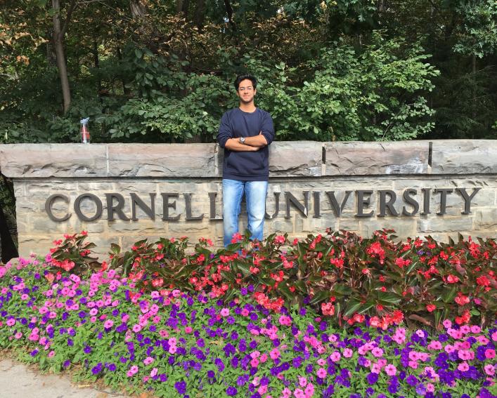 a college-aged man stands in a flower garden by a sign that reads Cornell University