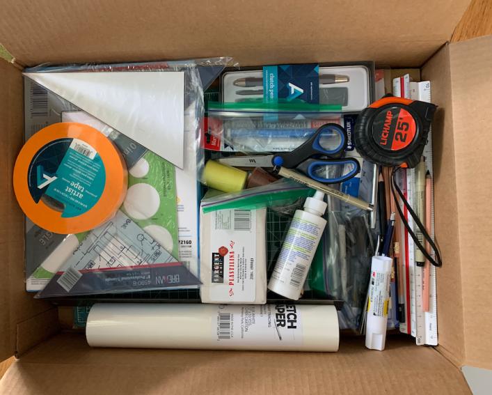 A cardboard box containing landscape architecture studio supplies, such as pencils, pens, charcoal sticks, erasers, engineering and architectural scales, drafting triangles, X-Acto knifes, cutting mats, sketchbooks and materials like paper, clay and cardboard.
