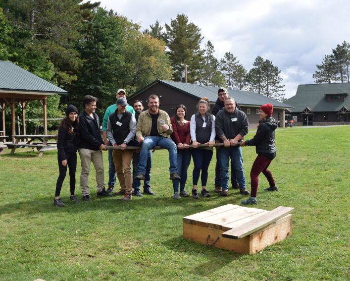 Group of students holds wooden plank with one student sitting on it
