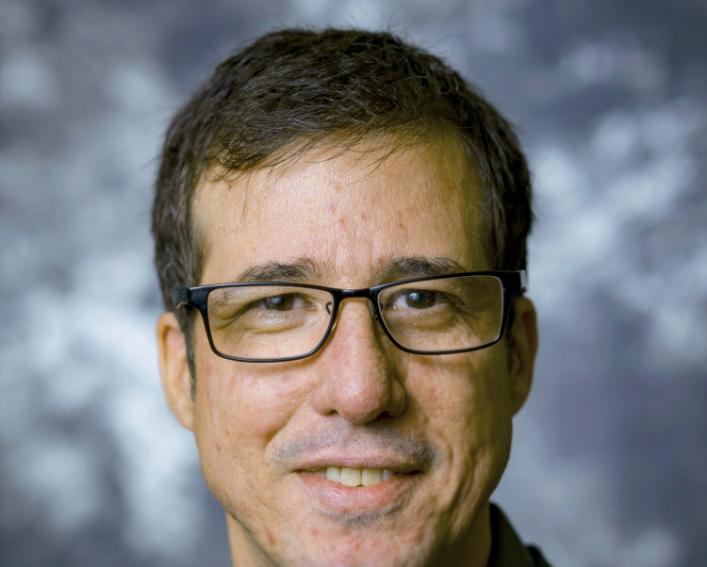 Headshot photograph of man in a dark blazer wearing glasses.