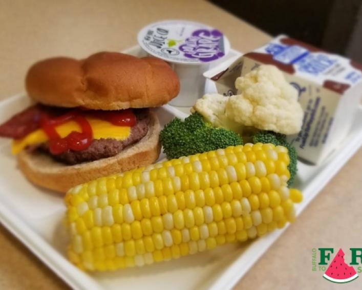 tray of food with half a cob of corn, hamburger, cauliflower, broccoli, grape juice cup, and chocolate milk carton