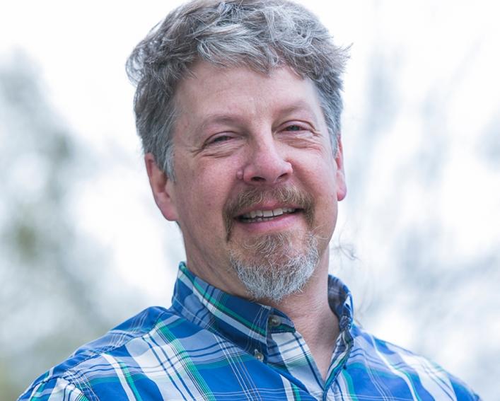 Headshot photograph of a man smiling wearing a blue plaid shirt.