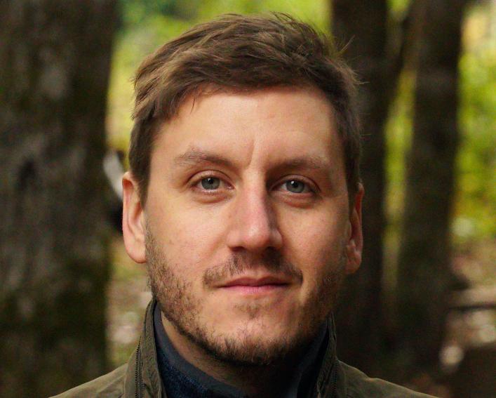 Headshot photograph of a man with short brown hair and stuble wearing a dark brown jacket.