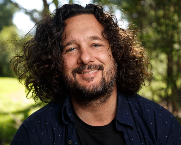 Headshot photograph of a man with shoulder length brown hair and a beard smiling wearing a black t shirt.