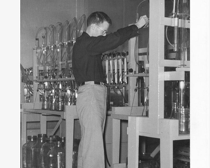 Black and white photo of a man in a lab with many different glass containers