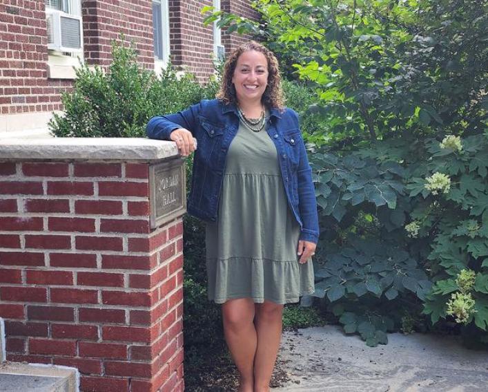 A woman in a green dress stands with one arm on a brick wall and greenery behind