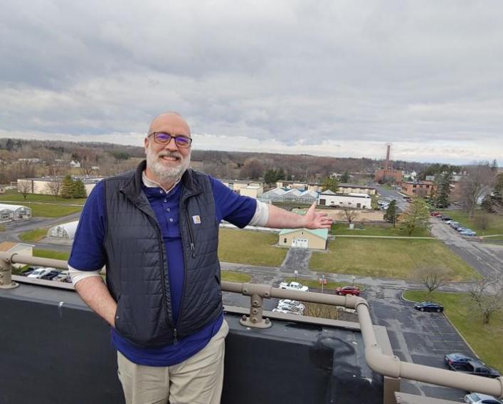 A man in glasses stands on a rooftop gesturing to a campus below