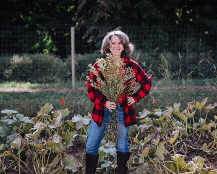 A woman in a red and black plaid shirt stands in a field with a bouquet of weeds