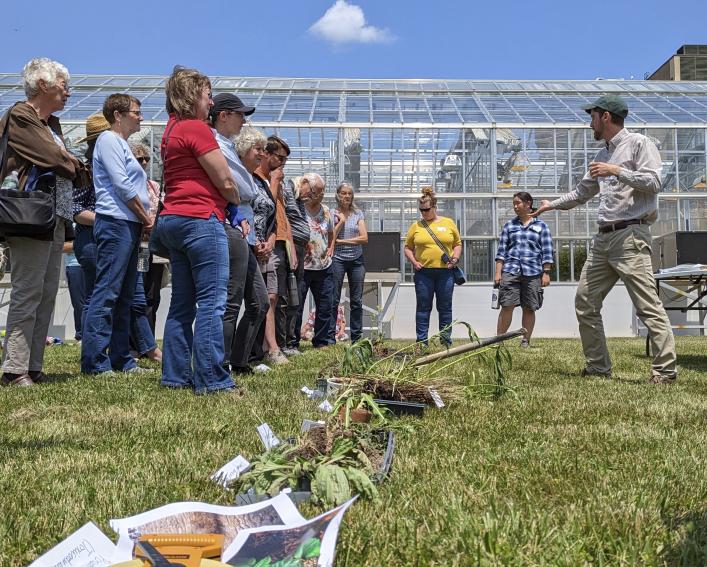 person talking to a group outdoors. A row of sample weeds on the ground.