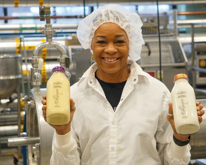 A woman wearing a white lab coat and hair net holding two bottles of milk. 
