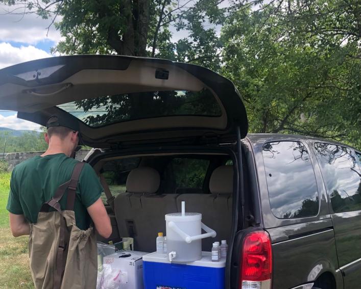 Photograph of a man wearing waders doing field water sampling out of the back of a minivan.