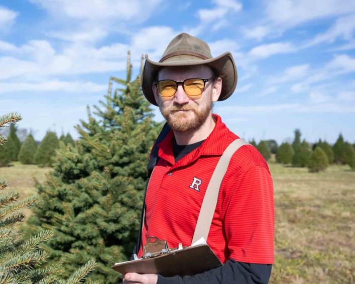 person with a Rutgers shirt, hat, glasses and clipboard standing in a Christmas Tree field.