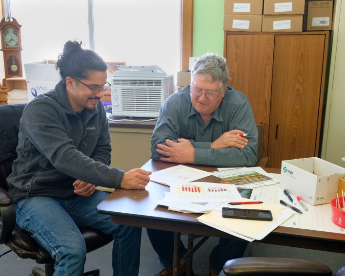 Two men sitting at a desk with papers on it talking.