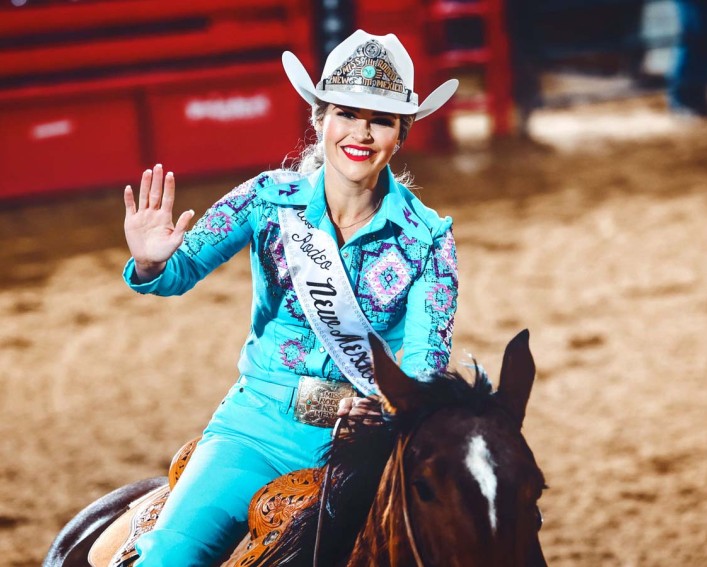 Person in a white cowboy hat and a blue pantsuit astride a brown horse