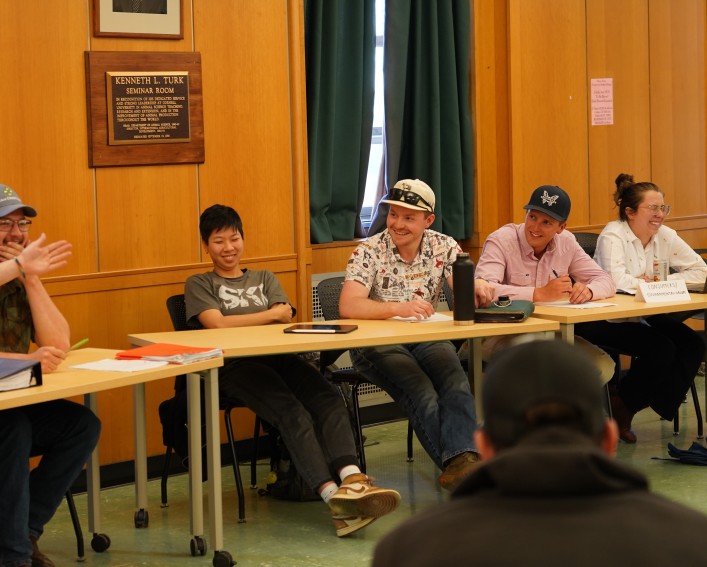 Five people at two separate tables gesturing towards one another smiling.