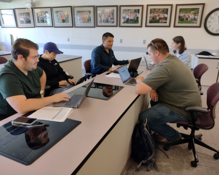 Five people sit around a table with their laptops having a discussion.