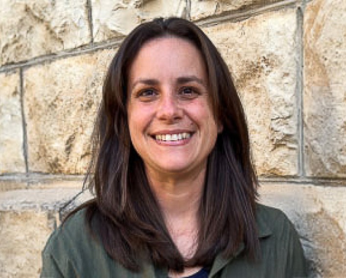 Headshot of Dr. Cassandra Olds, a person with long brown hair and a nice smile, wearing a green shirt and sitting in front of a tan stone wall.