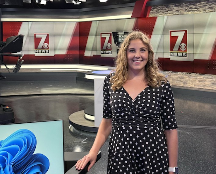 Person wearing a black polka-dot dress stands in a television news studio beside a desk, with cameras and ‘7 NEWS’ screens visible in the background.