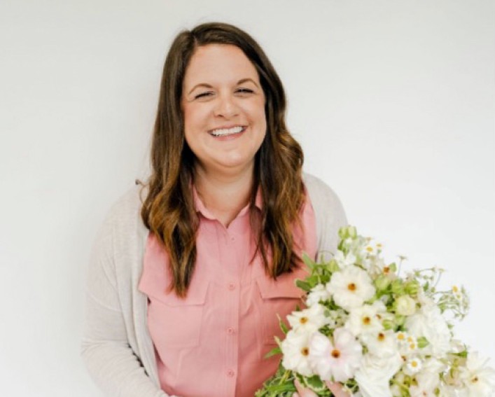 A portrait photo of a person with long brown hair smiling at the camera while holding a large bouquet of white flowers near their waist.