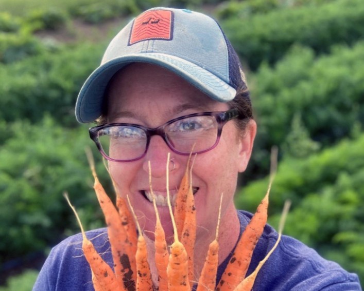 A person wearing eyeglasses and a baseball cap holds a large bunch of orange carrots in front of their face as they smile at the camera while standing in a lush carrot field.