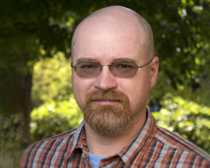 A headshot of a person with glasses and a light-colored goatee standing in front of a sun-dappled grove