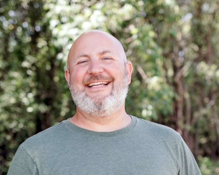 A person smiling at the camera while standing in a shady grove of trees and flowering bushes.
